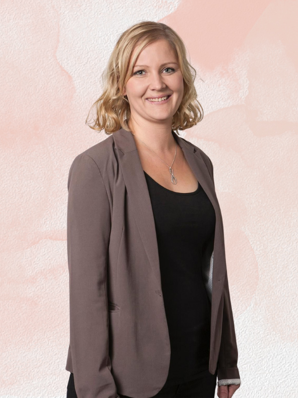 Nadine Warburton trägt ein schwarzes T-Shirt mit einem grauen Blaser. Sie hat blonde Haare und lächelt in die Kamera.